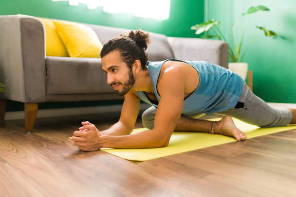 Vidám latin férfi gyakorolja a galamb pózt egy gyakorló szőnyegen a nappalijában. Vonzó férfi jógával gyakorol. - Fotó, kép