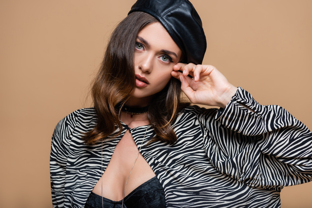 brunette woman in zebra print blazer and beret looking at camera while posing isolated on beige - Фото, зображення