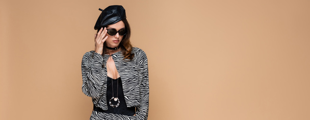 brunette woman in zebra print outfit and beret adjusting sunglasses isolated on beige, banner - Photo, Image