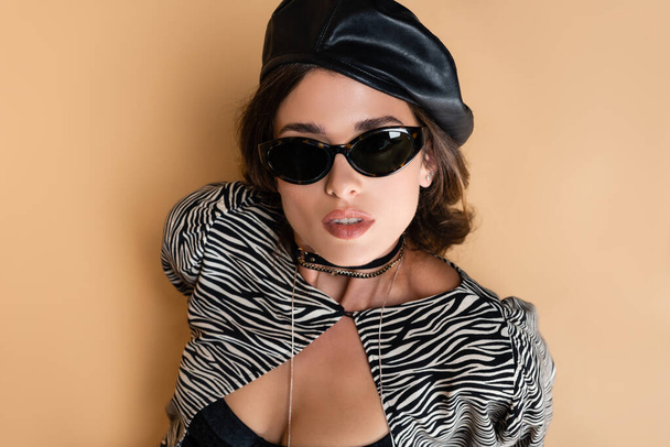 top view of brunette woman in zebra print outfit, leather beret and sunglasses looking at camera on beige  - Foto, Imagem