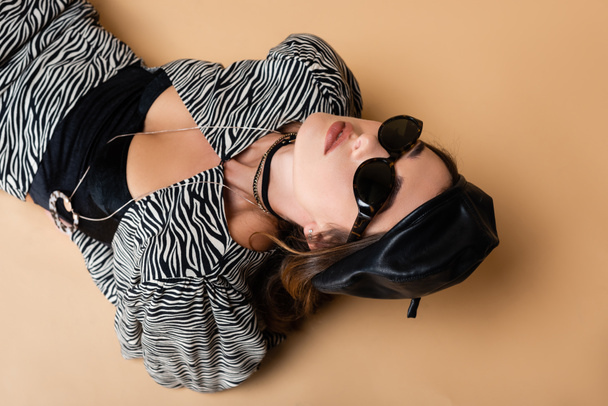 vista de ángulo alto de la mujer joven en traje de impresión de cebra, boina de cuero y gafas de sol posando en beige  - Foto, imagen