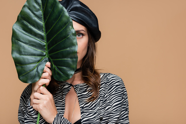 young woman in zebra print outfit and leather beret holding green leaf while covering face isolated on beige  - Foto, Bild
