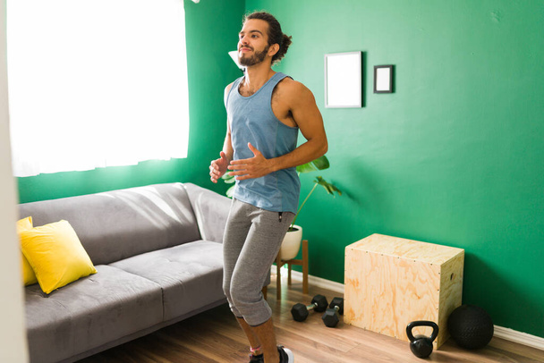 Genieten van een cardiotraining. Knappe latijnse man in actiefwear loopt op zijn plaats in de woonkamer - Foto, afbeelding