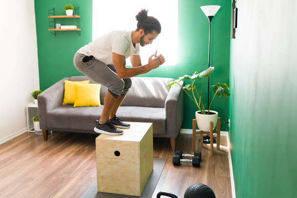 Залишатися здоровими. Активний іспаномовний чоловік стрибає на коробці плей під час перехресного тренування вдома
  - Фото, зображення