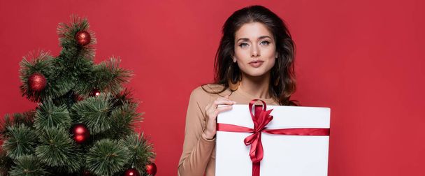 bella donna che tiene avvolto regalo vicino all'albero di Natale isolato sul rosso, banner - Foto, immagini
