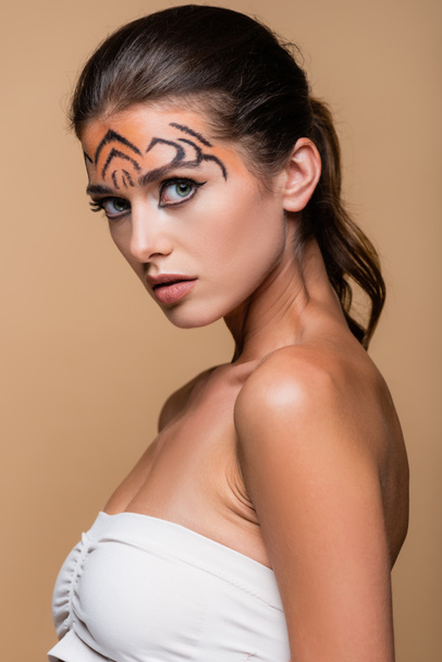 pretty woman with bare shoulders and tiger makeup looking at camera isolated on beige - Foto, imagen