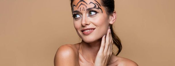 wesoła kobieta z gołymi ramionami i makijażem tygrysa odwracając wzrok odizolowana na beżu, sztandar - Zdjęcie, obraz