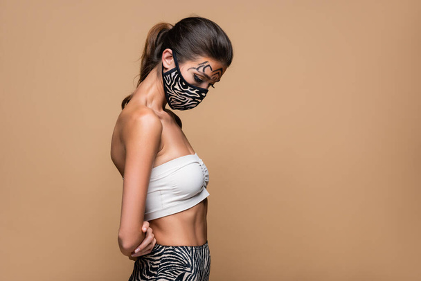 mujer con maquillaje de tigre y máscara protectora animal print mirando hacia abajo aislado en beige - Foto, imagen