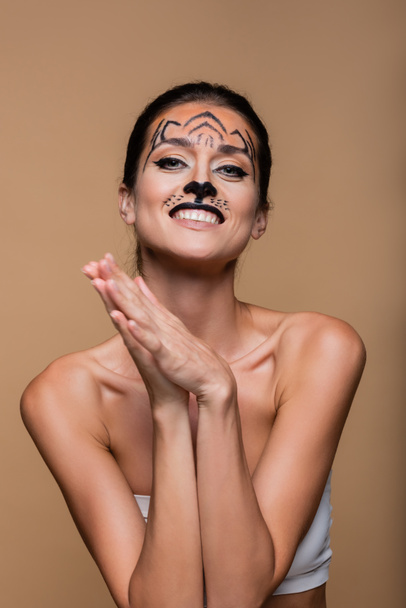 χαμογελαστή γυναίκα με γυμνούς ώμους και μακιγιάζ τίγρης που ποζάρει απομονωμένη σε μπεζ - Φωτογραφία, εικόνα