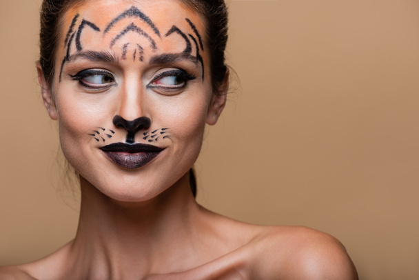 sly woman with bare shoulders and tiger makeup looking away isolated on beige - Photo, Image