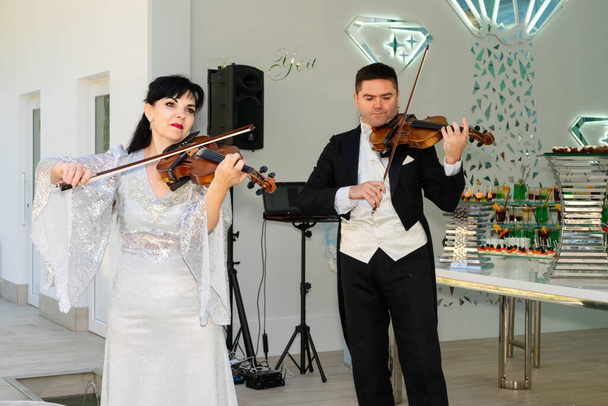 夜の衣装を着た男と女がバイオリンを弾く。ヴァイオリニストのデュオ. - 写真・画像