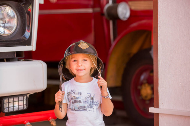 Child, cute boy, dressed in fire fighers cloths in a fire station with fire truck, childs dream - Fotoğraf, Görsel