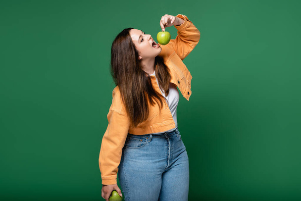 Young plus size woman holding apple near open mouth isolated on green  - Photo, Image