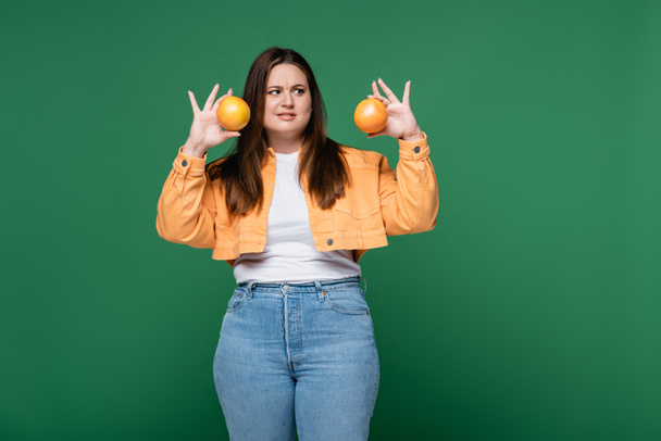 Confused woman with overweight holding oranges isolated on green  - Valokuva, kuva