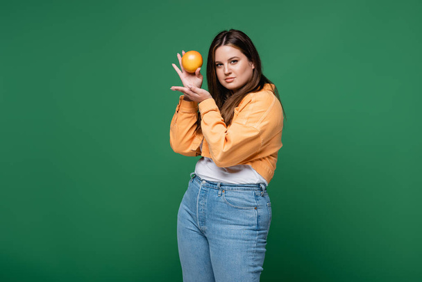 Pretty woman with overweight holding orange and looking at camera isolated on green  - 写真・画像