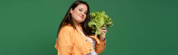 Uśmiechnięta kobieta z nadwagą trzymająca sałatę i patrząca na kamerę odizolowaną na zielono, baner - Zdjęcie, obraz