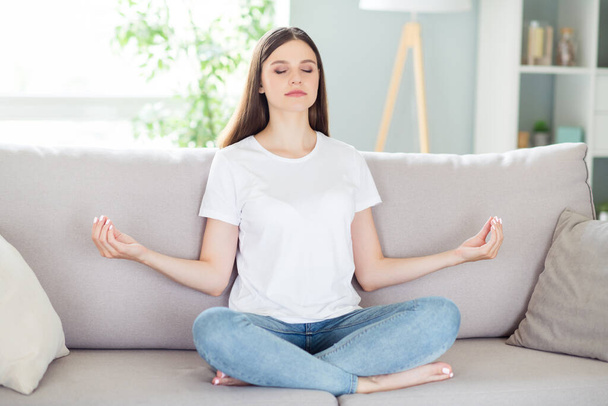 Portrait of attractive calm focused girl sitting on divan in lotus pose meditating resting at home indoors - Φωτογραφία, εικόνα