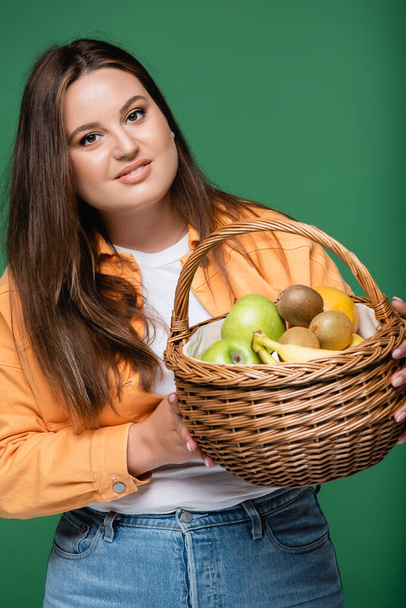 Mujer morena con cesta de sobrepeso con frutas aisladas en verde  - Foto, Imagen