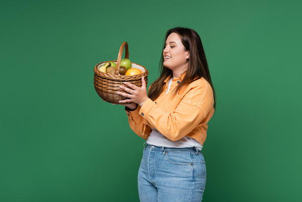 Mujer positiva con cesta de sobrepeso con frutas frescas aisladas en verde  - Foto, Imagen