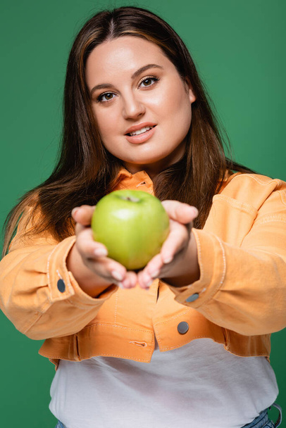Pretty woman with overweight holding blurred apple and looking at camera isolated on green  - Photo, Image