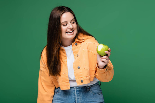 Cheerful body positive woman looking at apple isolated on green  - Fotoğraf, Görsel