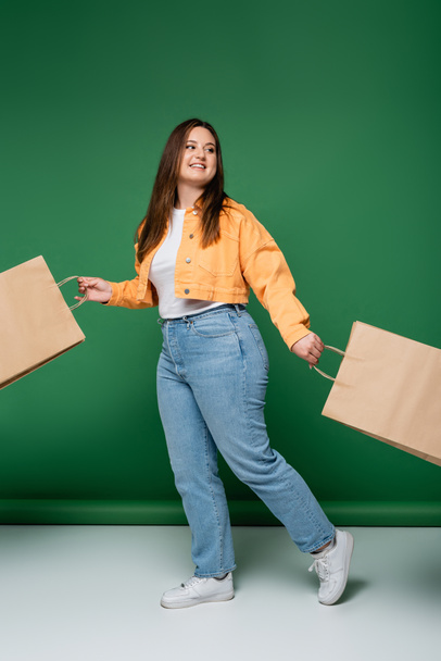 Cheerful body positive woman with craft shopping bags walking on green background - Foto, afbeelding