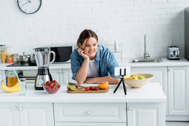 Uśmiechnięta kobieta z nadwagą patrząc na smartfona w pobliżu owoców i blendera na stole kuchennym  - Zdjęcie, obraz