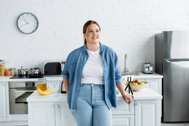 Donna felice con sovrappeso in piedi vicino al tavolo della cucina con cellulare e cibo  - Foto, immagini