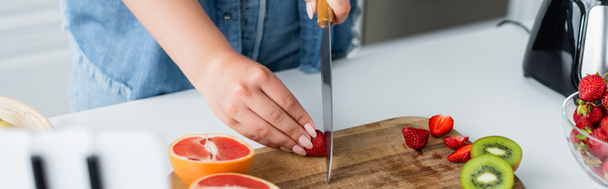 Cropped view of woman cutting fruits near blurred cellphone in kitchen, banner  - Foto, afbeelding