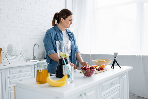 Vue latérale de plus taille femme coupe des fruits mûrs près du smartphone et mélangeur à la maison  - Photo, image