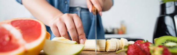 Cropped view of woman cutting banana near blurred fruits in kitchen, banner  - Фото, изображение