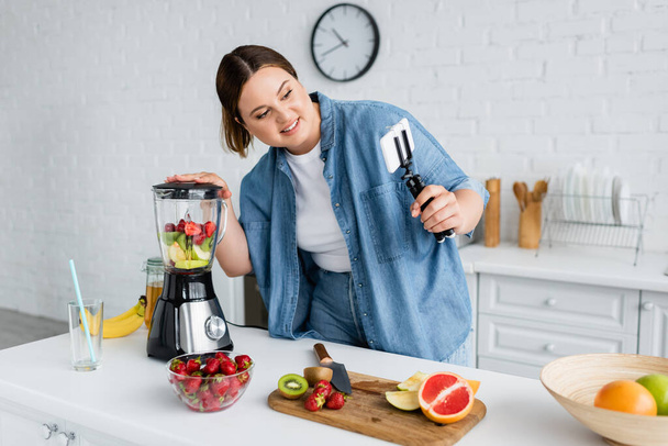 Cheerful plus size blogger using smartphone while preparing smoothie in kitchen  - Foto, afbeelding