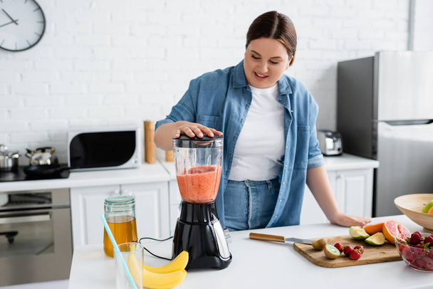 Smiling plus size woman preparing smoothie in blender near honey and fresh fruits in kitchen  - Photo, Image