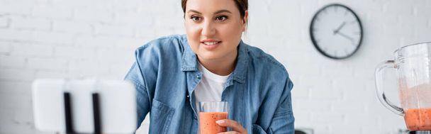 Sonriendo más tamaño mujer sosteniendo smoothie cerca borrosa teléfono celular en la cocina, pancarta  - Foto, Imagen