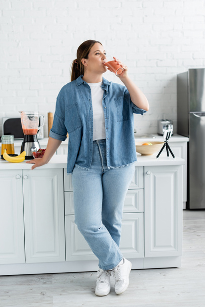 Mladá žena s nadváhou pití smoothie v blízkosti mobilního telefonu a zralé ovoce v kuchyni  - Fotografie, Obrázek
