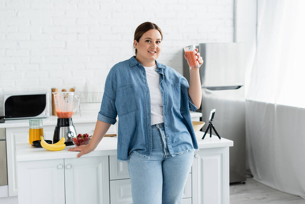 Улыбающаяся женщина plus size держит стакан смузи и смотрит на камеру возле мобильного телефона на кухне  - Фото, изображение
