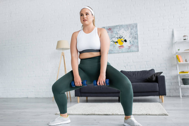 Plus size woman doing squat while training with dumbbells at home  - Valokuva, kuva