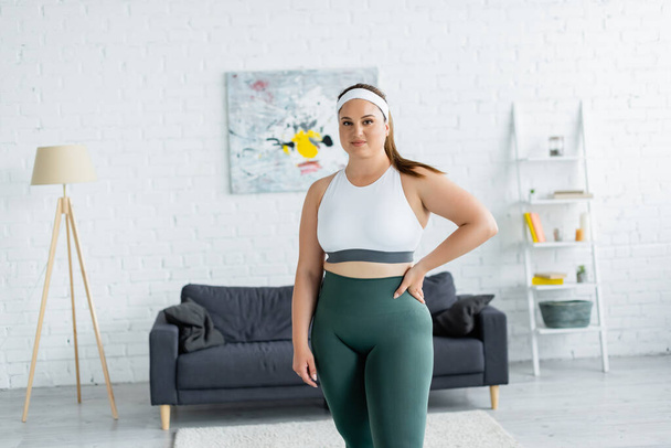Mosolygó sportos nő túlsúlyos fogja a kezét csípő a nappaliban  - Fotó, kép