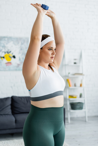 Young plus size sportswoman exercising with dumbbell in living room  - Foto, afbeelding