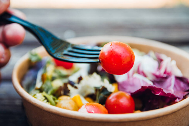 Close-up de uma mão com um garfo e um tomate. No fundo uma salada mediterrânea em um recipiente de papelão. - Foto, Imagem