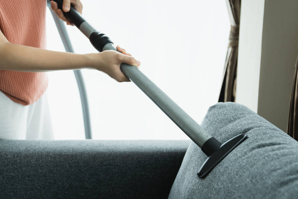 Asian housewife using a wireless vacuum machine to clean a sofa in living room close up. Asian housekeeper vacuuming in living room. Chores and daily housework activities concept. - Фото, изображение
