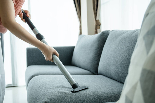 Asian housewife using a wireless vacuum machine to clean a sofa in living room close up. Asian housekeeper vacuuming in living room. Chores and daily housework activities concept. - Foto, Imagen