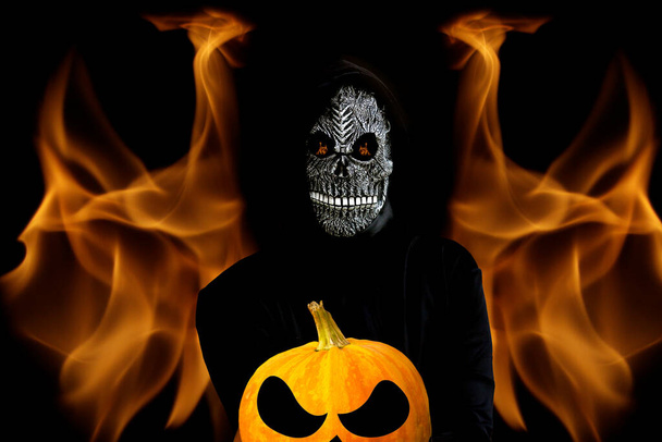 Grande faucheuse tenant la tête de citrouille halloween. L'homme dans le masque de la mort avec la flamme du feu dans les yeux et sourire sur fond de flamme noir foncé de feu de l'enfer. Concentration sélective. - Photo, image