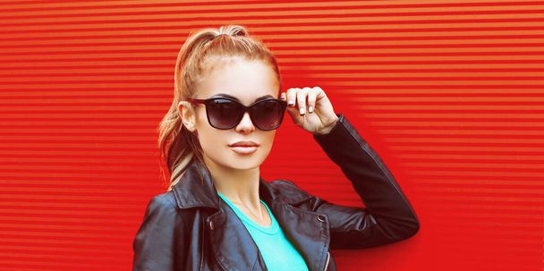 Stylový portrét krásné blondýny na sobě černé rockové bundy a sluneční brýle na červeném pozadí - Fotografie, Obrázek