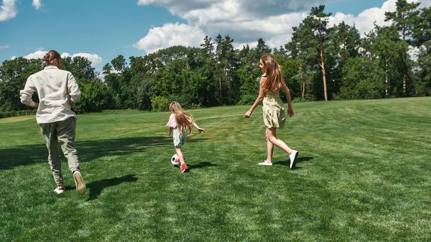 Ενεργός νεαρή οικογένεια παίζει ποδόσφαιρο μαζί με τη μικρή κόρη στο γρασίδι στο πάρκο σε μια καλοκαιρινή μέρα - Φωτογραφία, εικόνα