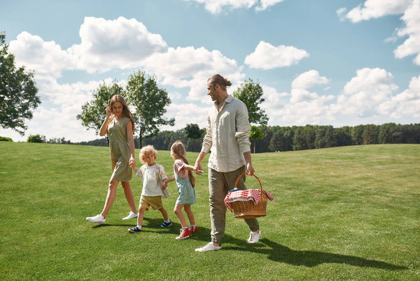 Full length shot of happy young parents with two little kids walking in green park, ready for picnic in nature on a summer day - Zdjęcie, obraz