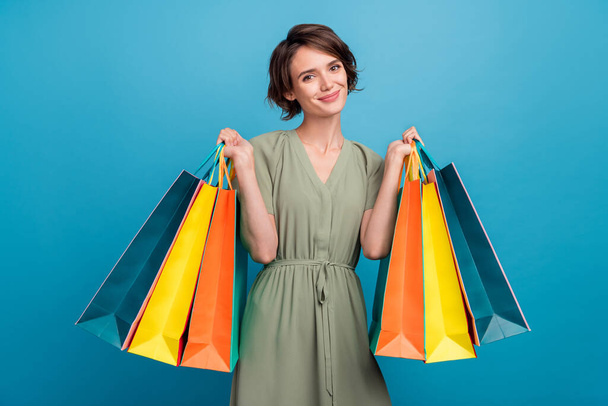 Photo of cute young brunette lady hold bags wear khaki dress isolated on blue color background - Φωτογραφία, εικόνα