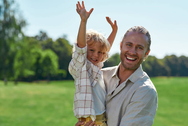 Excited father smiling at camera, holding his child while playing with him on grass field on a summer day. Little boy raised his arms, looking cheerful - Foto, Bild