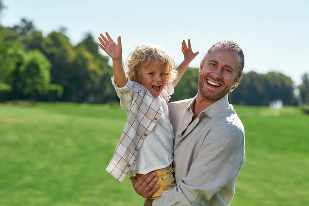 Loving young dad smiling at camera, holding his child while playing with him on grass field on a summer day. Little boy raised his arms, looking cheerful - Фото, изображение