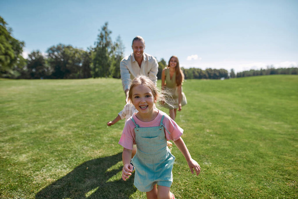Joyful cute little girl looking excited at camera, having fun together with her parents and brother, running in green park on a summer day - 写真・画像